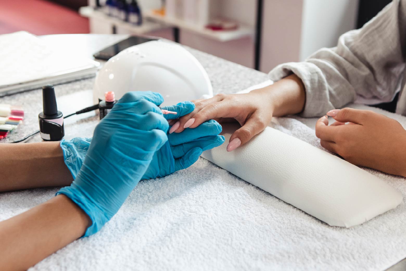 nail artist wearing blue latex gloves painting client’s nails on top of a white pillow and towel in a nail salon